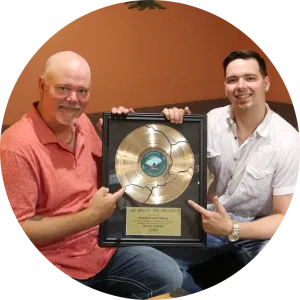 Two men sitting together, smiling and pointing at a framed gold record award